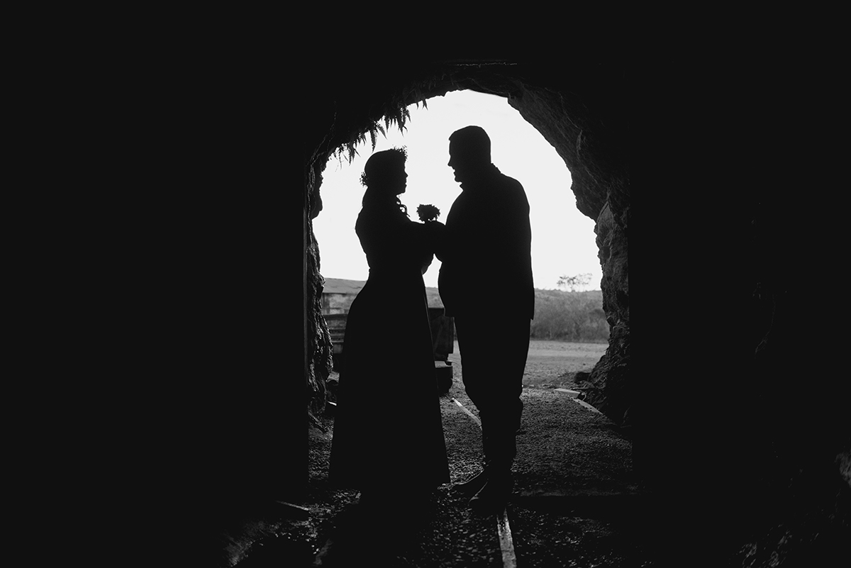 Foto contra a luz em preto e branco do casal de noivos na entrada de um tunel ferroviário nas minas do camaquã durante ensaio do casal em seu pré wedding.