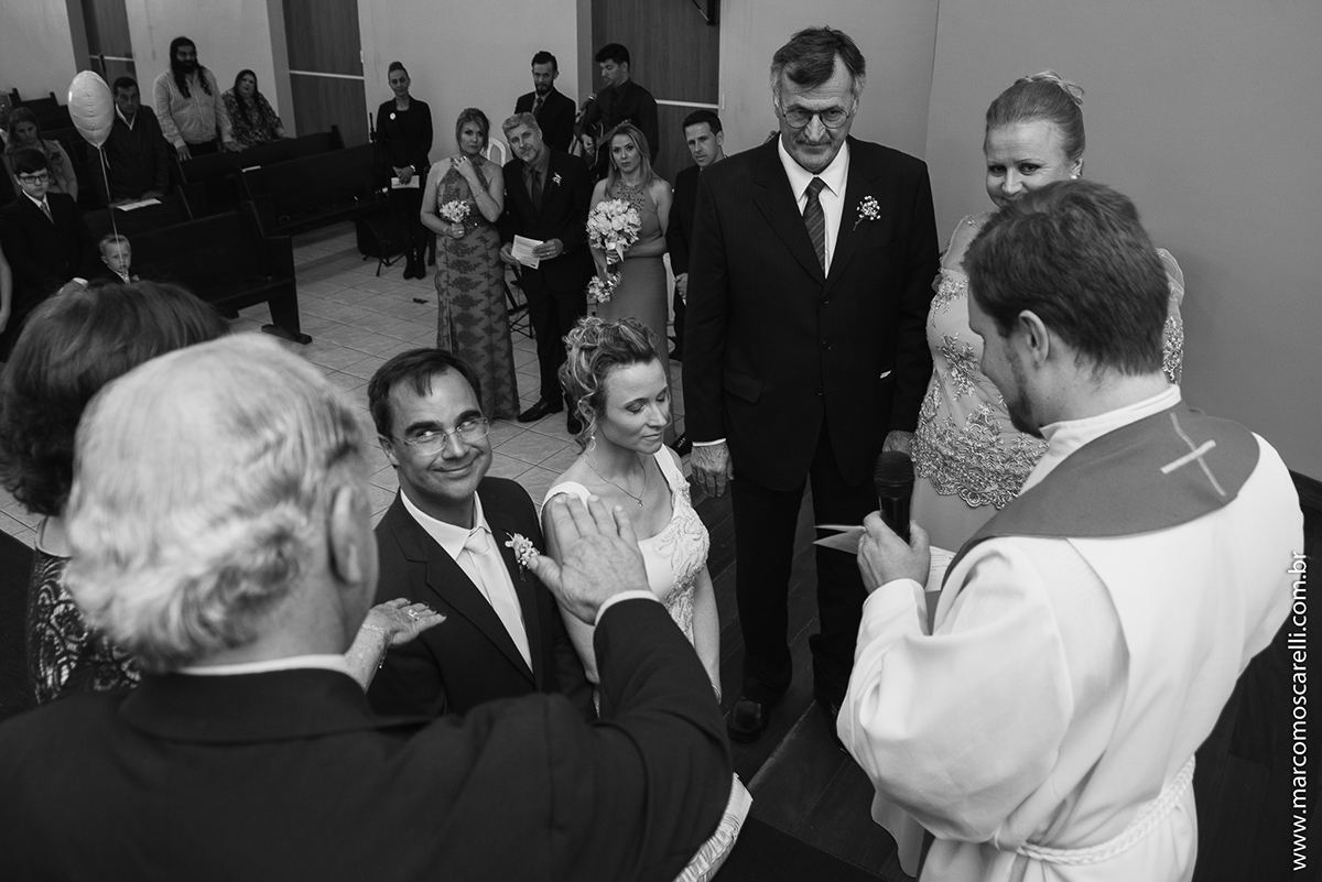 Foto em preto e branco dos noivos ajoelhados no altar da igreja e o noivo dando olhar engração para a sua ma. Foto por Marco Moscarelli