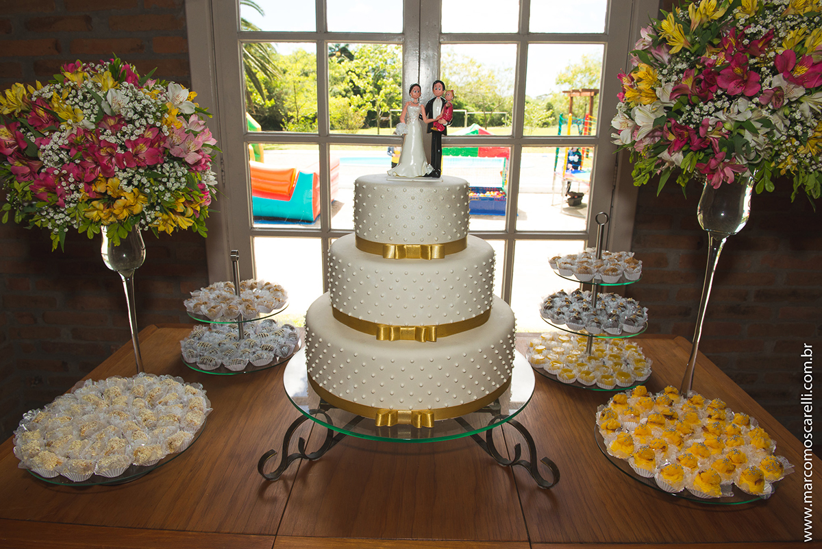 Mesa do bolo linda decorada com flores, does e um lindo casal de noivos no topo do bolo. Foto por Marco Moscarelli