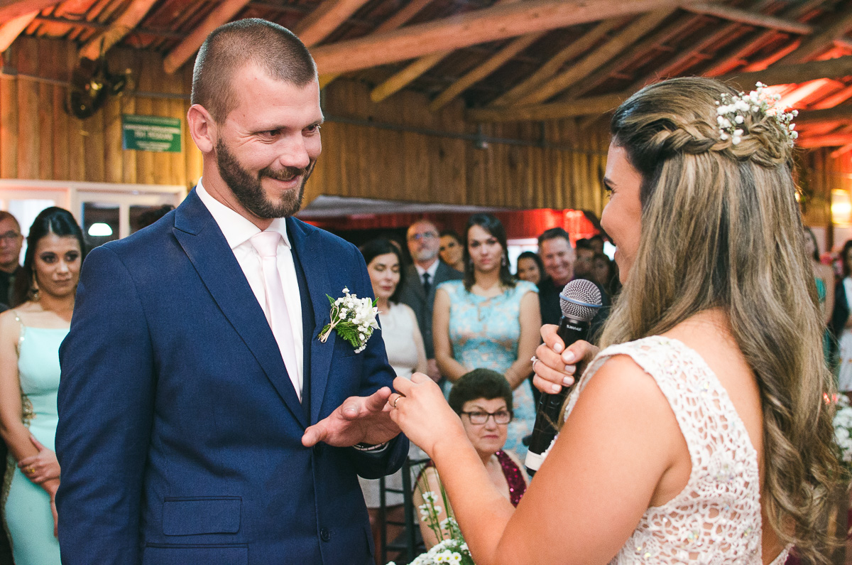 Noiva colocando a aliança no dedo do noivo sorridente. Foto Marco Moscarelli. Casamento Clube Caça e Pesca Pelotas