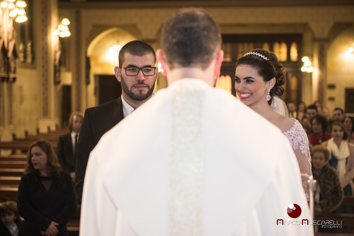 Noiva sorrindo para o noivo no altar da Paroquia São Pedro enquanto padre celebra o casamento. Foto por Marco Moscarelli