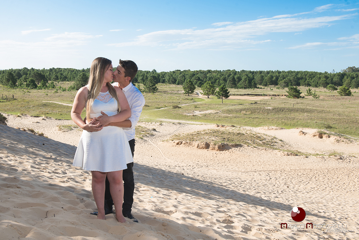 Casal de noivos posando abraçados no alto de uma duna de areia com um lindo bosque de acácias ao fundo em ensaio pré-wedding. Foto por Marco Moscarelli