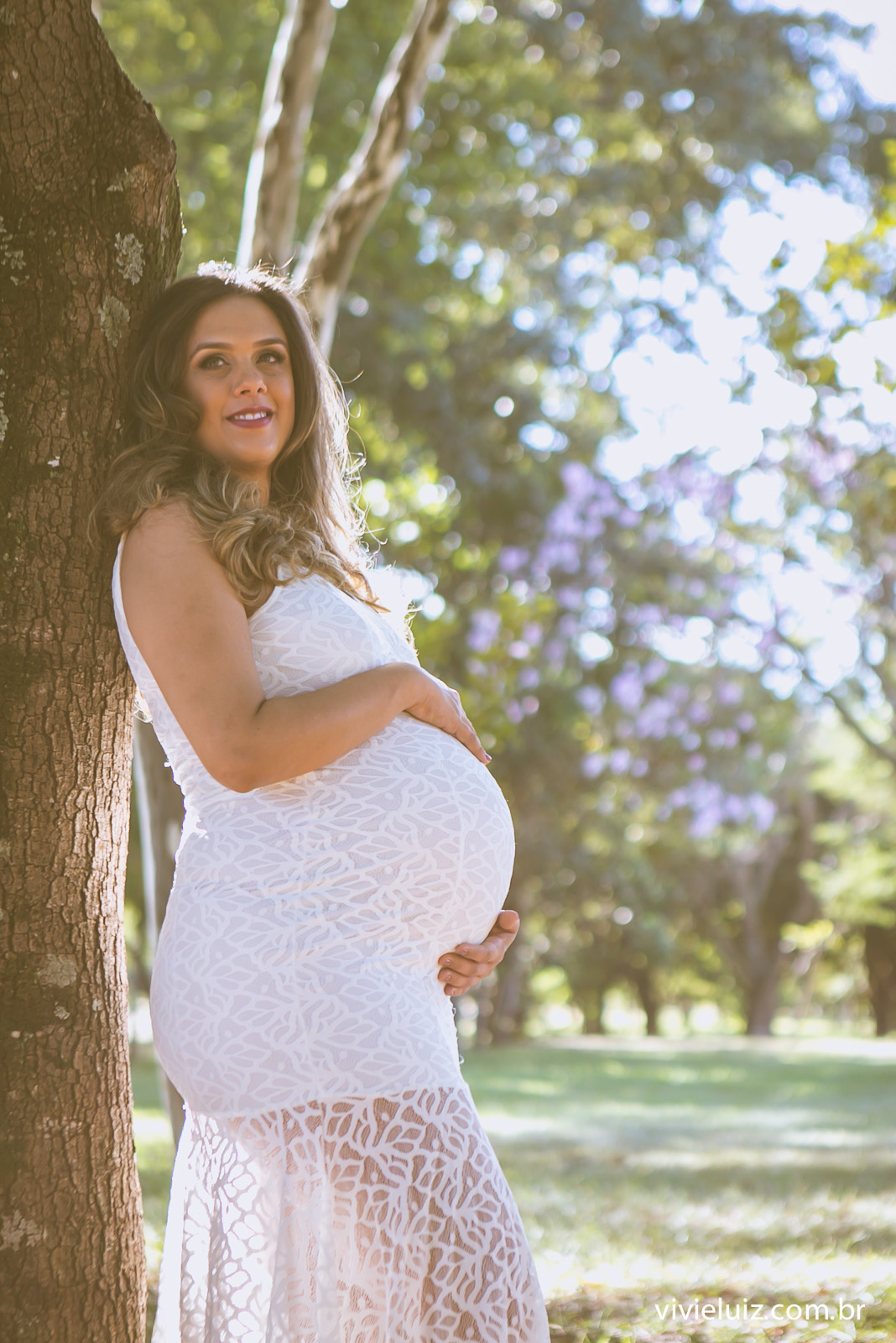Mãe com um vestido branco gestante encostada na árvore
