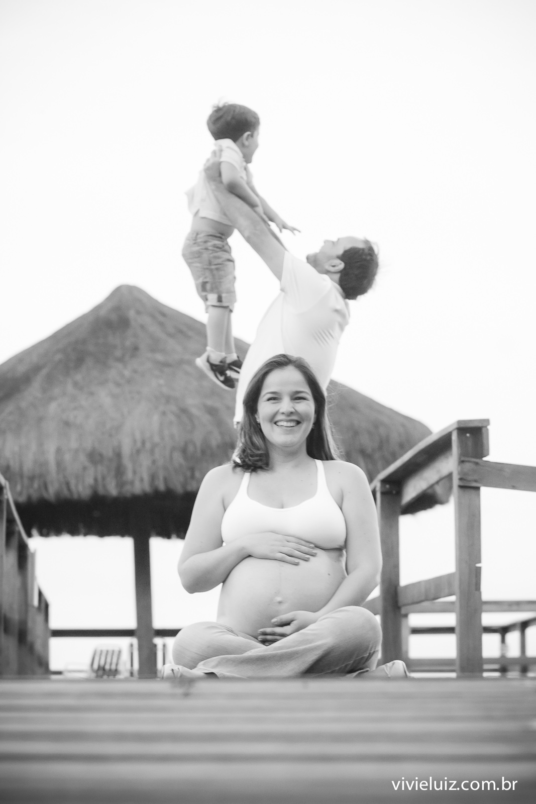 Mãe grávida sentada E pai ao fundo jogando o filho mais velho para cima