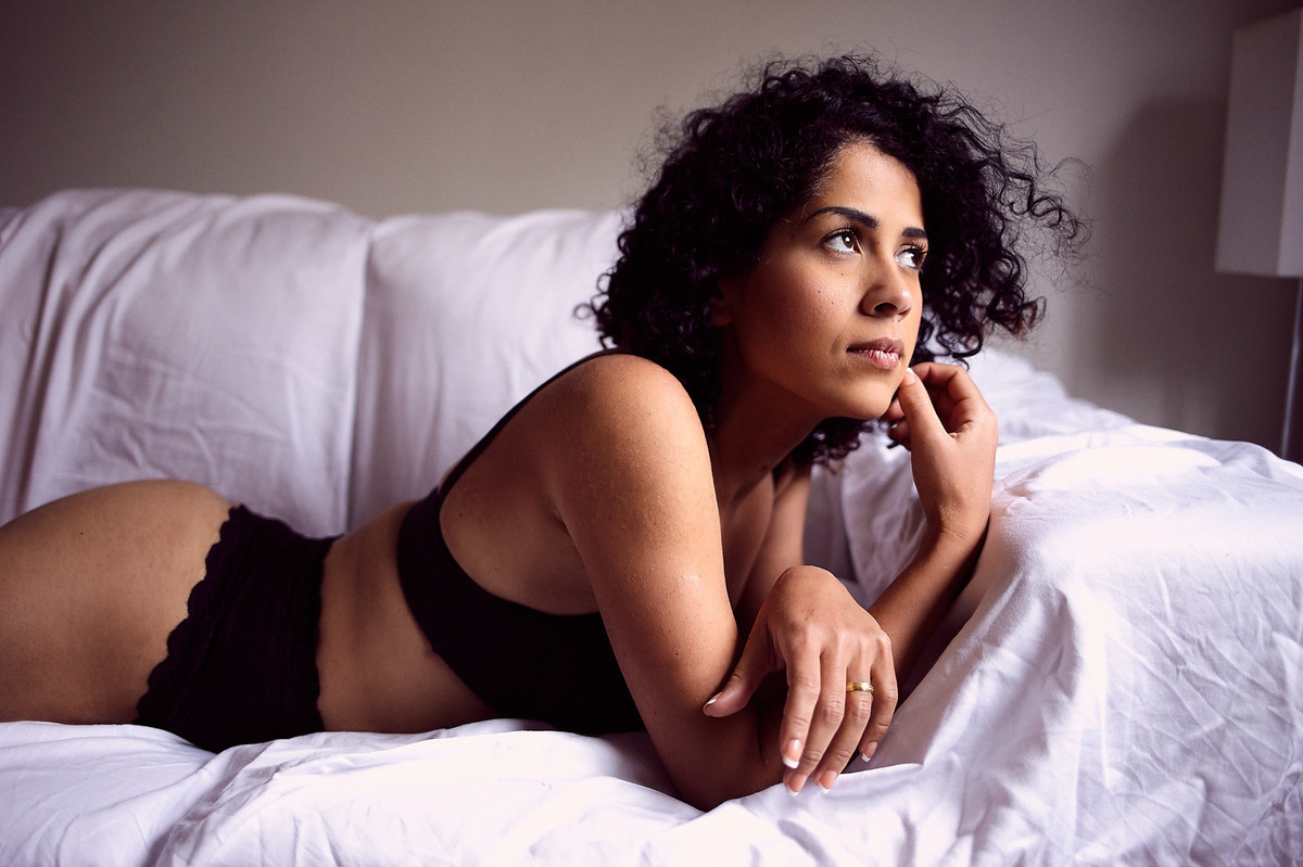 Deitada sobre o sofá, usando lingerie preta, Adriely olha serenamente em direção à luz, com seus cabelos movimentados pelo vento, em foto de Jon K. Roz, retratista especializado em ensaios femininos