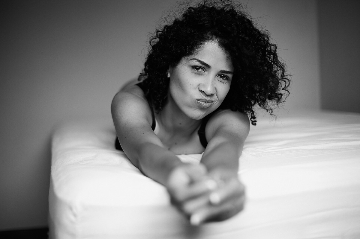 Charme, ao fazer careta, da linda Adriely, deitada sobre a cama, com os braços estendidos em direção à câmera, em click preto e branco do fotógrafo Jon K. Roz, que faz retratos femininos em Curitiba