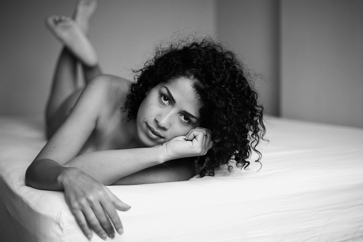 Click, em preto e branco, do fotógrafo Jon K. Roz, de retratos intimistas femininos, com a bela Adriely, sobre a cama, deitada, com o corpo inclinado para a direita, olhando diretamente