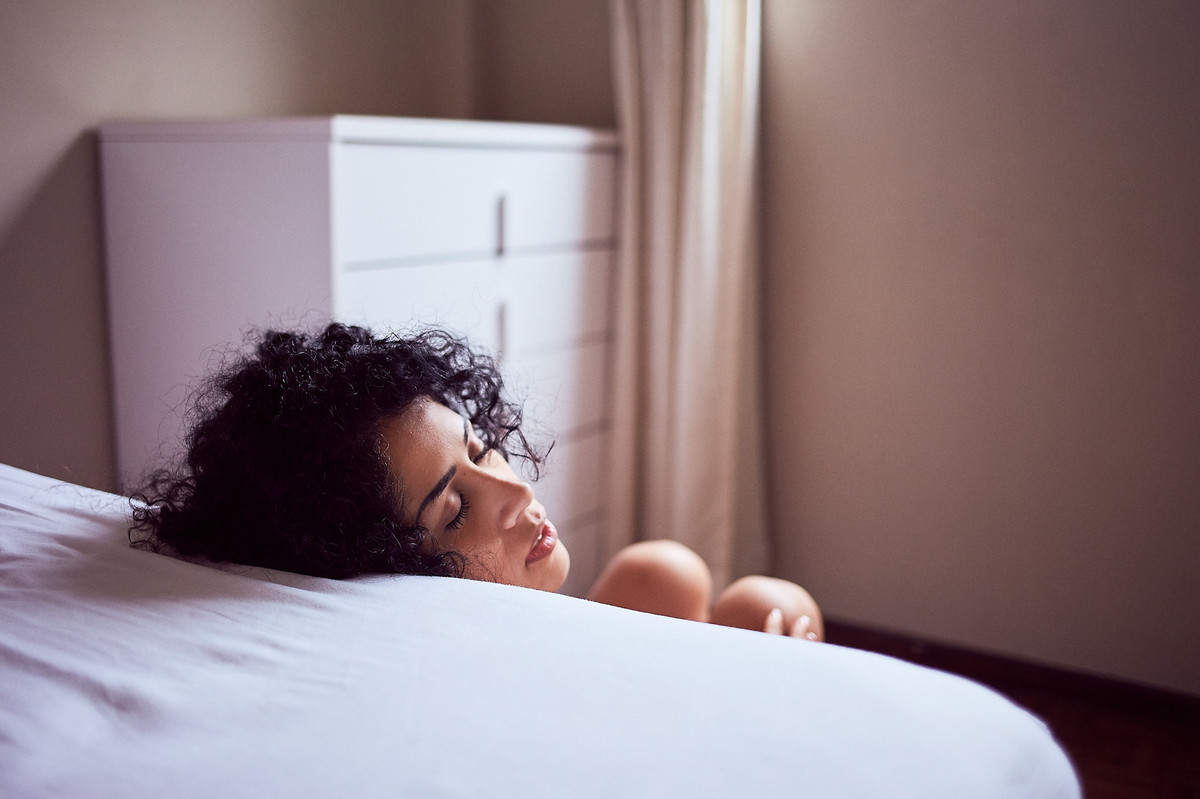 Adriely, sentada ao pé da cama, no chão, olhos fechados e cabeça apoiada, em ensaio boudoir do fotógrafo Jon K. Roz, em mais um belo retrato feminino