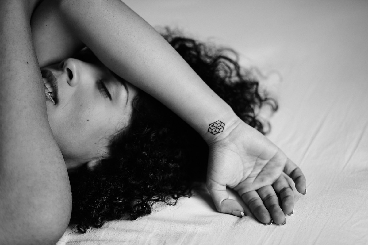 A bela Adriely, deitada sobre a cama com olhos fechados e braço sobre a cabeça, revelando tatuagem no pulso, em click preto e branco do fotógrafo Jon K. Roz, de Curitiba