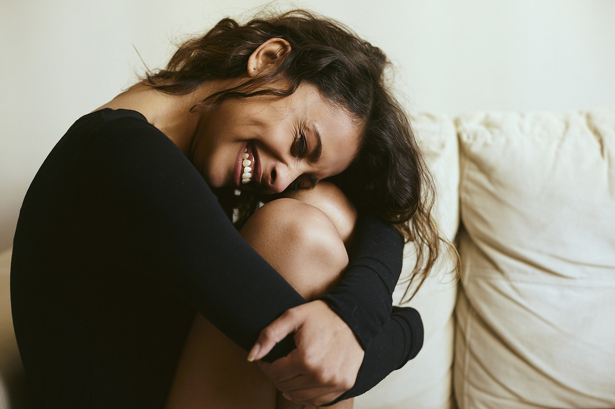 A linda Andry, sorrindo sentada no sofá, abraçando as próprias pernas, em ensaio boudoir do fotógrafo Jon K. Roz, especializado em retratos femininos