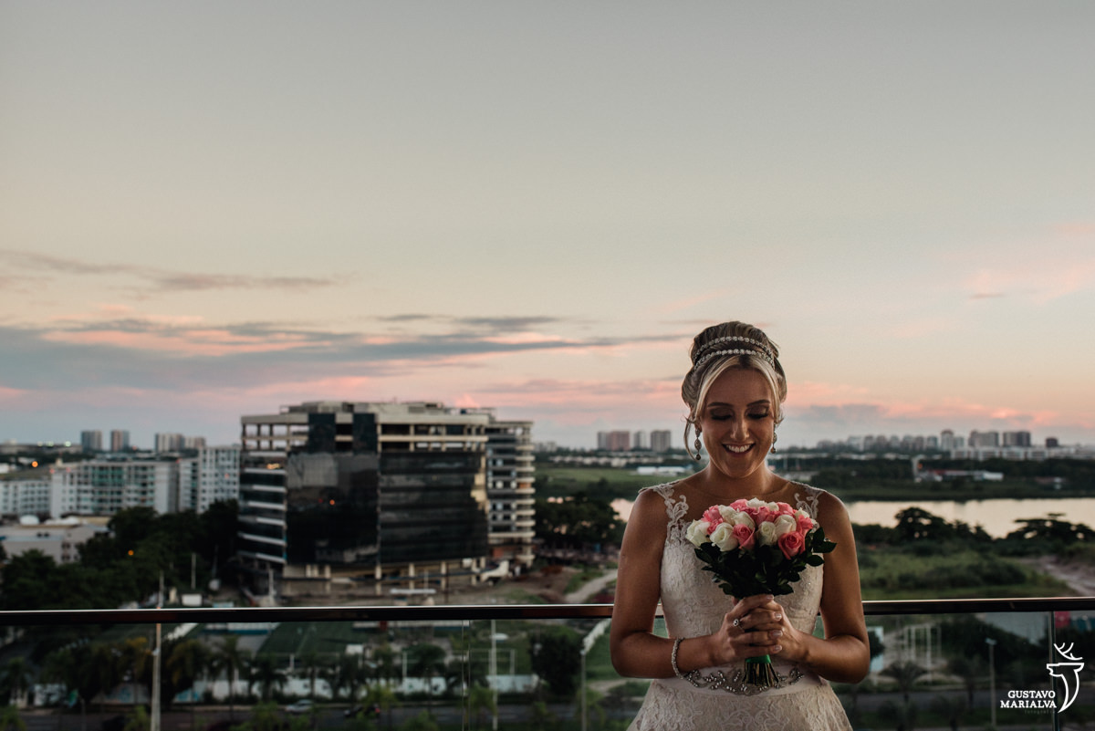 noiva segurando o buque e sorrindo com a barra da tijuca ao fundo, no por do sol, durante o making of do casamento no hotel hilton no rio de janeiro