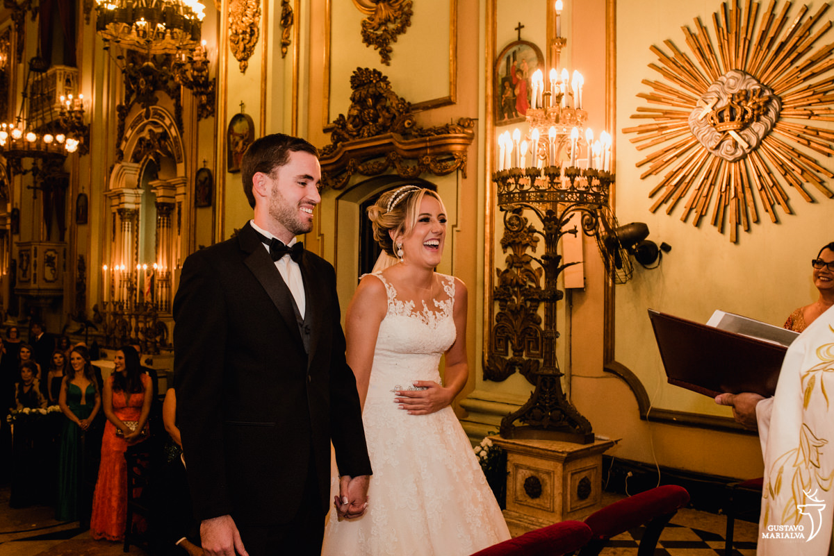 noiva e noivo sorrindo, enquanto a noiva coloca a mão na barriga, na cerimônia de casamento na igreja são josé no rio de janeiro