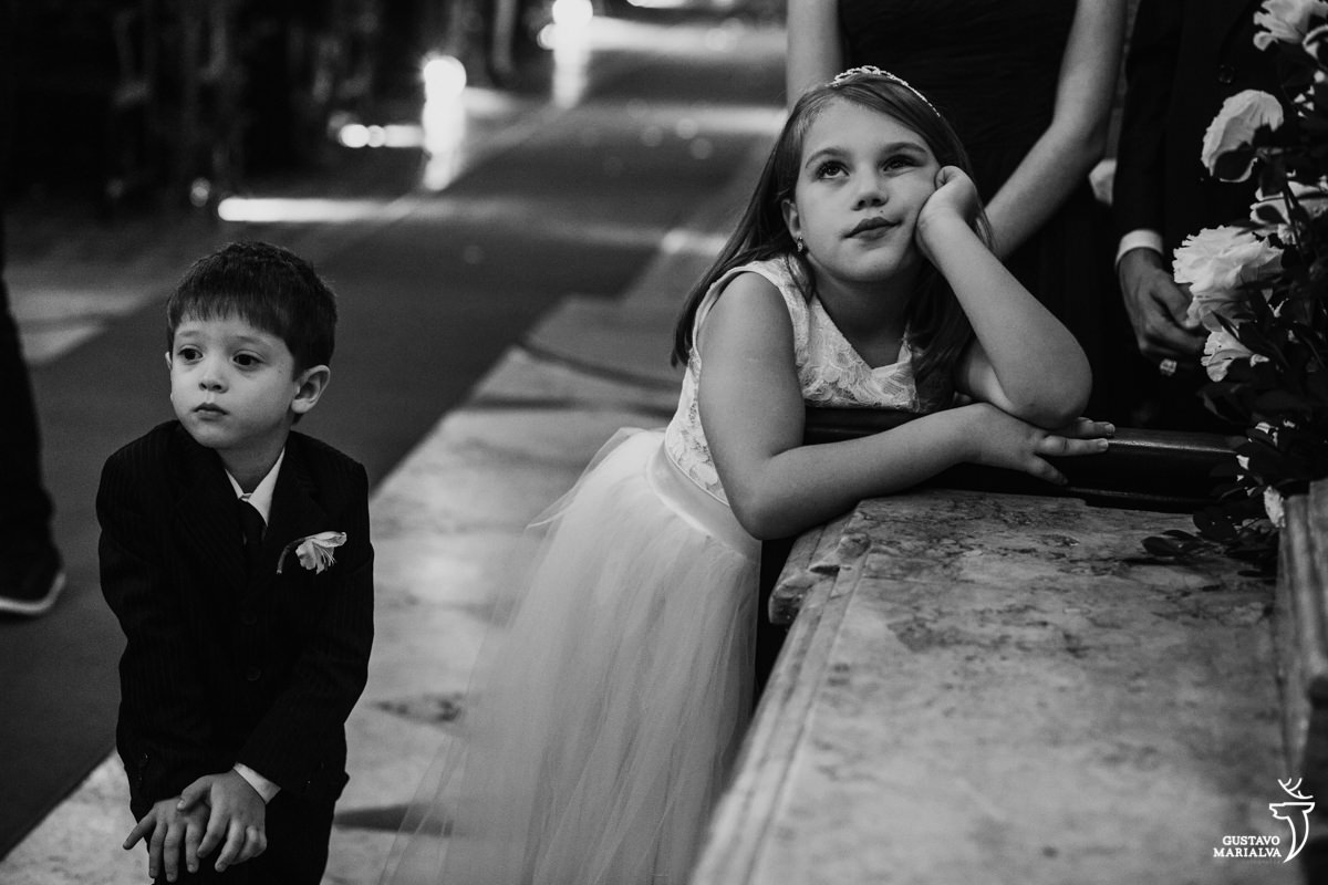 daminha apoiando a mão no rosto com sono enquanto pajem presta atenção na cerimônia de casamento na igreja são josé no rio de janeiro