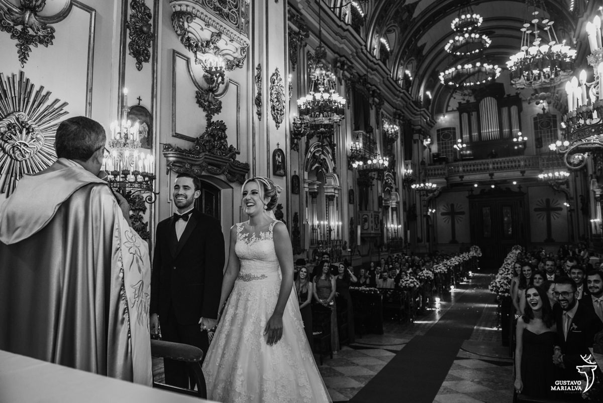 noivos sorrindo enquanto escutam o padre falar e convidados sorrindo ao fundo durante a cerimônia de casamento na igreja são josé no rio de janeiro