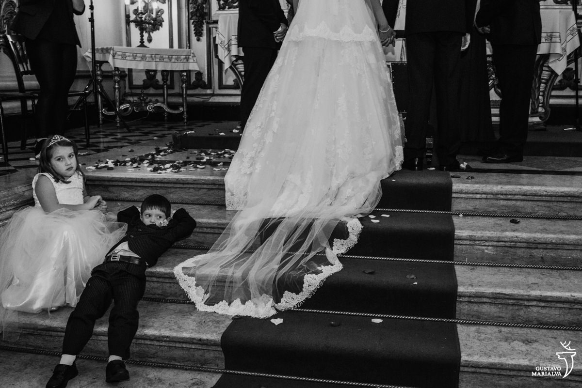 pajem deitado na escada do altar, com a daminha sorrindo enquanto os noivos observam o padre falar durante a cerimônia de casamento na igreja são josé no rio de janeiro