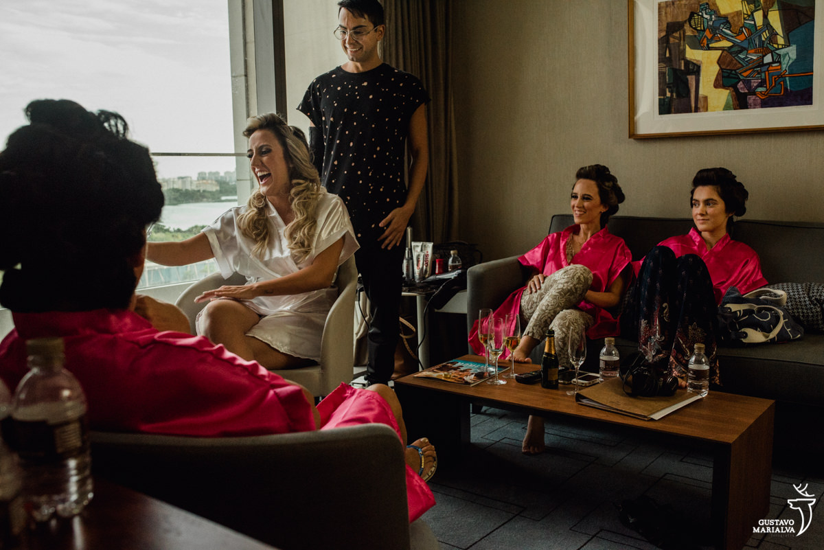 mãe observando a noiva sorrir enquanto o maquiador daniel wagner arruma o seu cabelo e irmã e madrinha sorrindo ao fundo no making of do casamento no hotel hilton no rio de janeiro