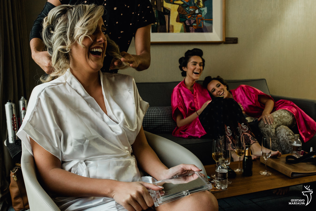 noiva gargalhando e irmã e madrinha se abraçando no fundo durante o making of do casamento no hotel hilton no rio de janeiro