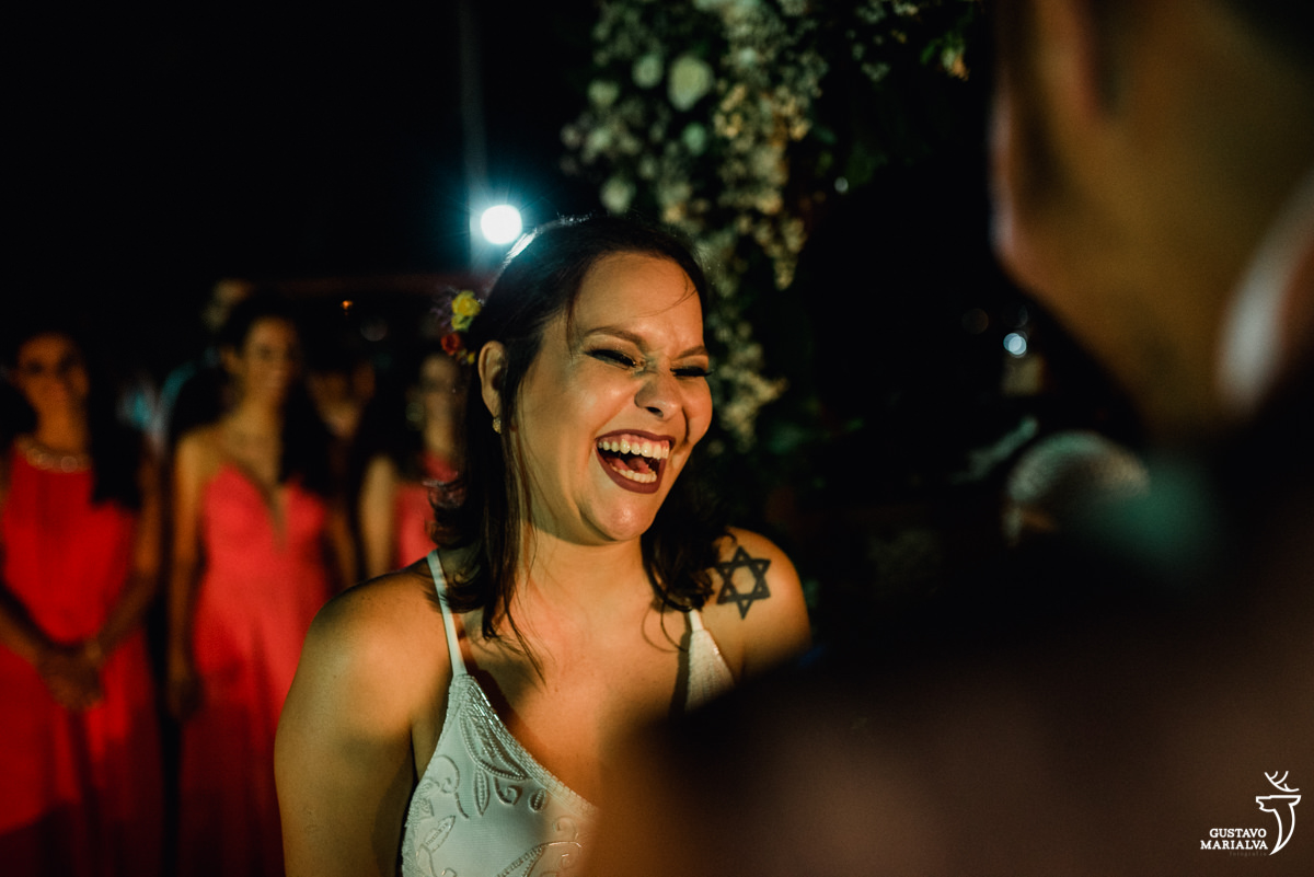 noiva sorri emocionada com as madrinhas ao fundo enquanto o noivo lê os votos na cerimônia de casamento no Uniq Beach Lounge em Búzios