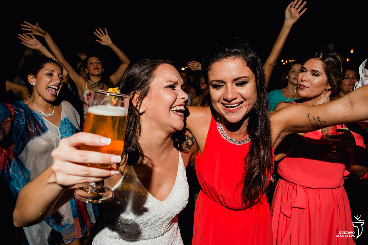 noiva com copo de cerveja na mão abraça a madrinha emocionada enquanto convidados dançam no fundo