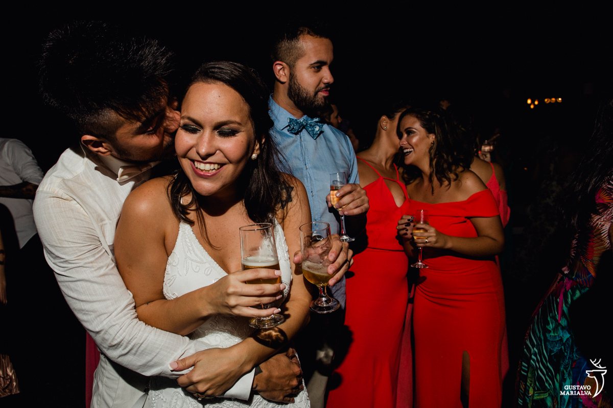 noivo abraça noiva por trás e dá um beijo enquanto madrinhas conversam sorrindo ao fundo no Uniq Beach Lounge em Búzios
