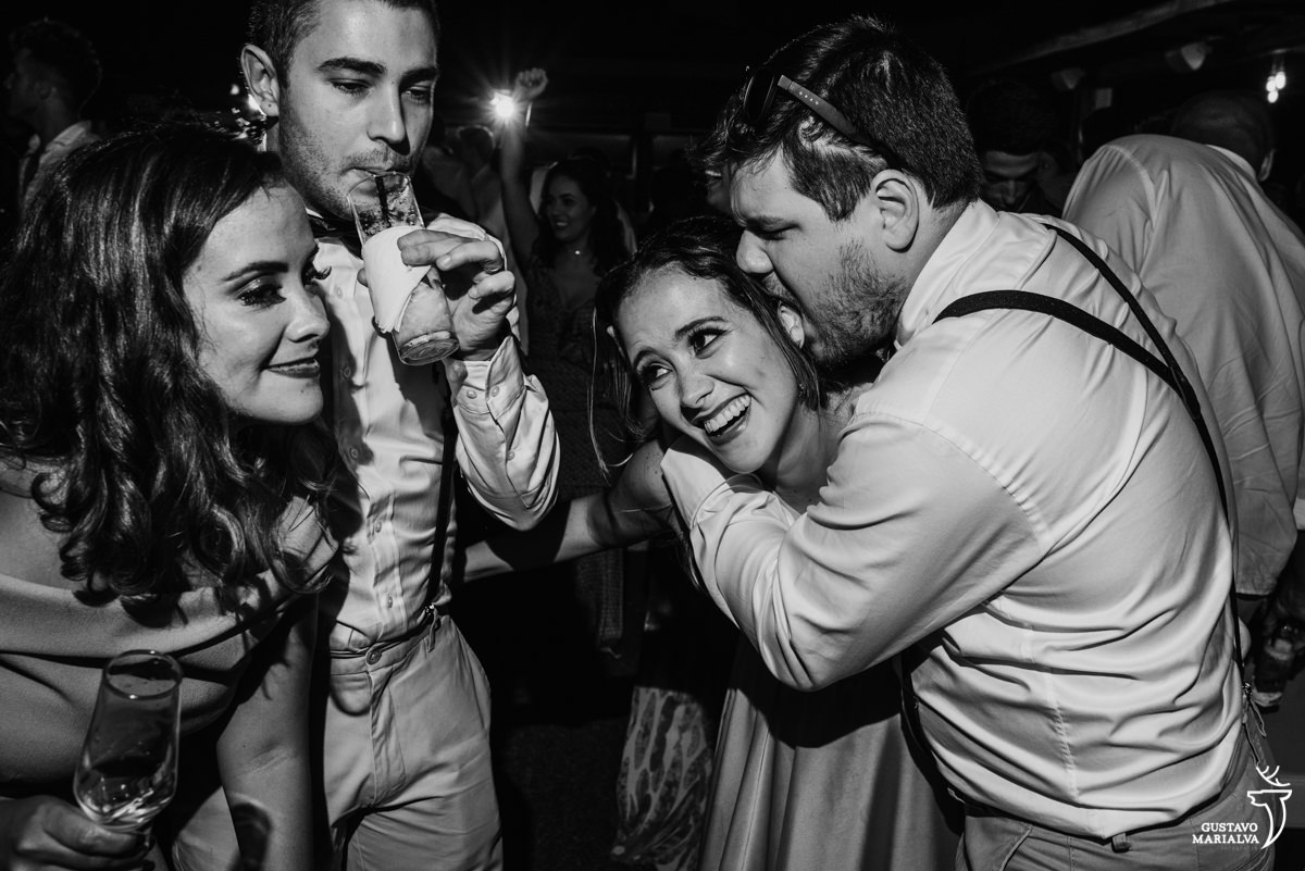 padrinho morde a orelha da madrinha enquanto amigo toma um drink e outra madrinha dança sorrindo