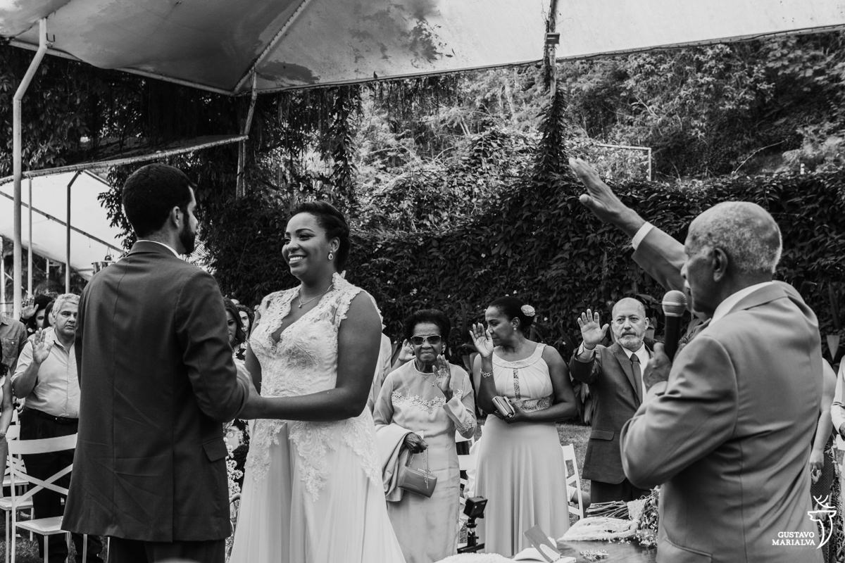 avô da noiva abençoa o casal com o braço estendido com a noiva sorrindo e mãe da noiva emocionada no fundo