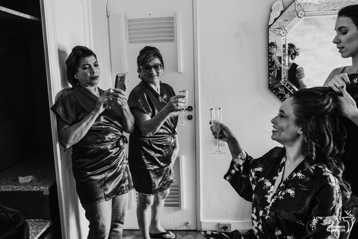 noiva sorri para mãe que tira foto com celular enquanto é observada pela sogra no making of do casamento