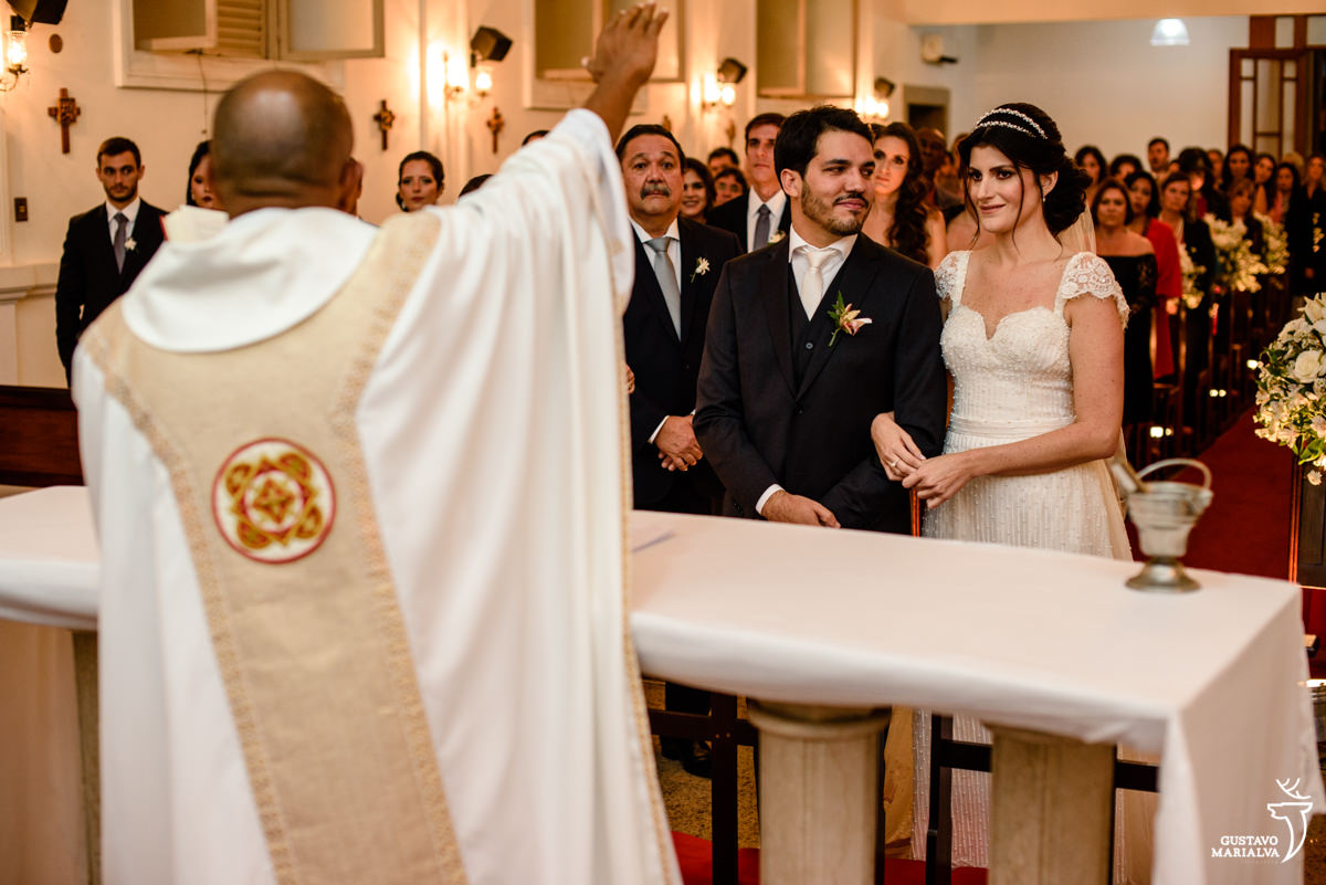 noivo olhando para noiva com olhar apaixonado enquanto são abençoados pelo padre durante a cerimonia do casamento