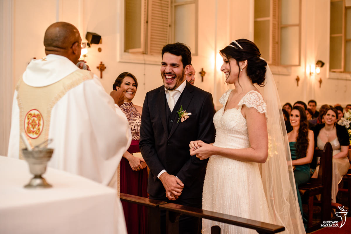 noivo sorrindo com o sermão do padre enquanto é observado pela noiva
