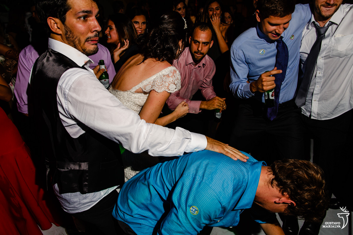 noivo faz careta ao dança com amigo agachado na sua frente enquanto casal de amigos se abraça ao fundo