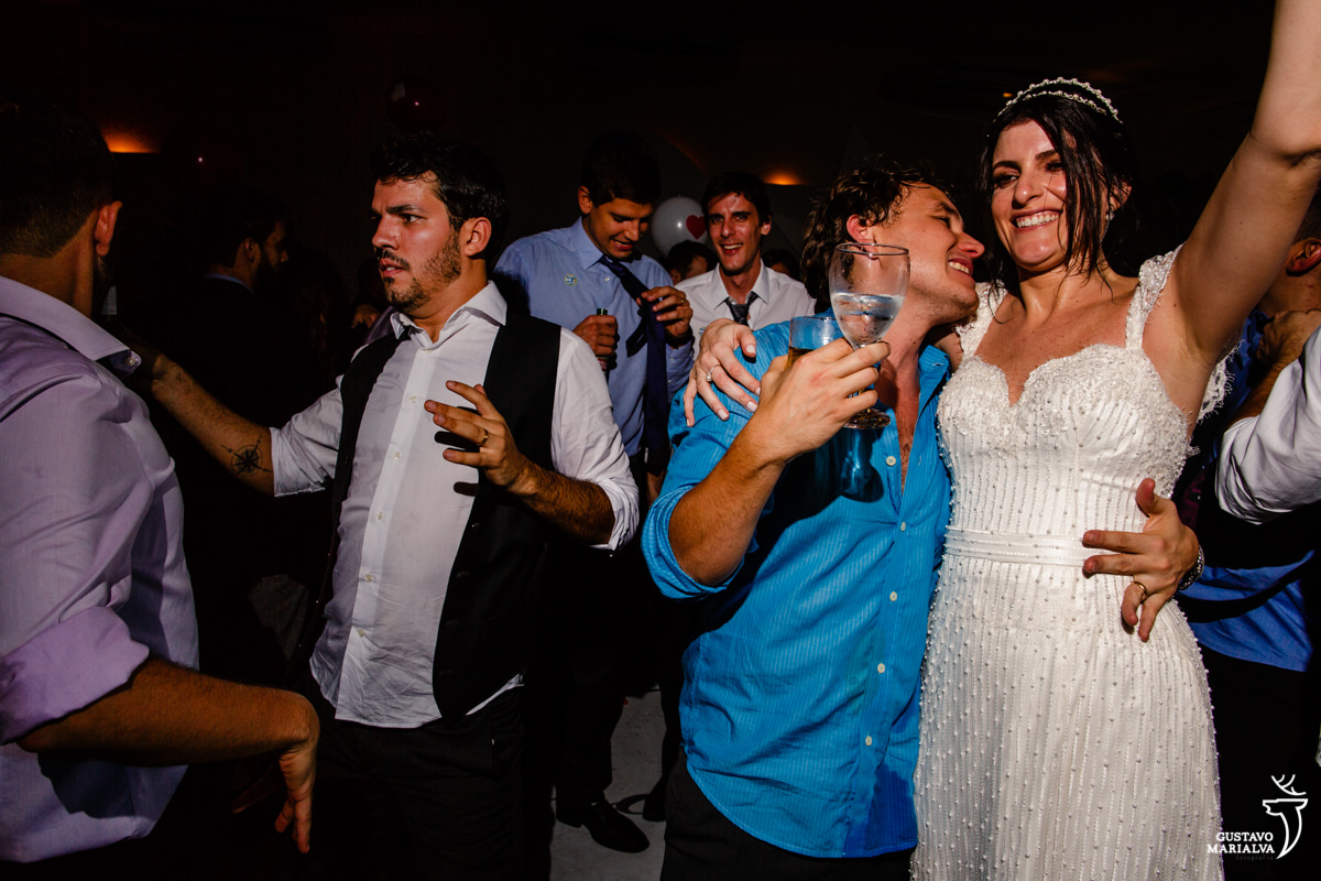 noiva abraça amigo com taça de espumante nas mãos enquanto noivo dança com amigo