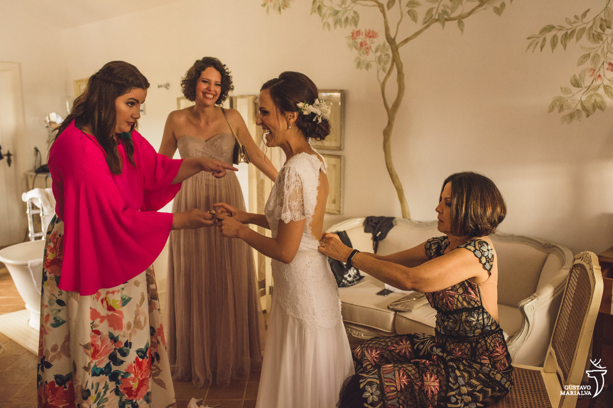 mãe da noiva fecha o vestido da noiva enquanto as amigas observam emocionadas e sorridentes