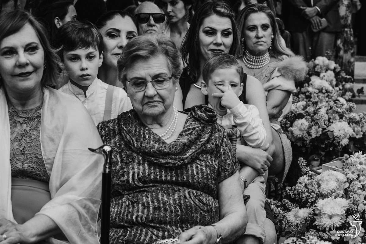 menino boceja com a mão no rosto enquanto senhora observa a cerimônia