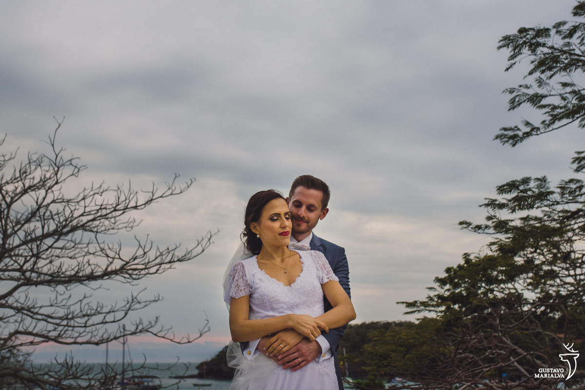 noivo abraça a noiva por trás durante retrato de casamento com a praia dos ossos em búzios ao fundo