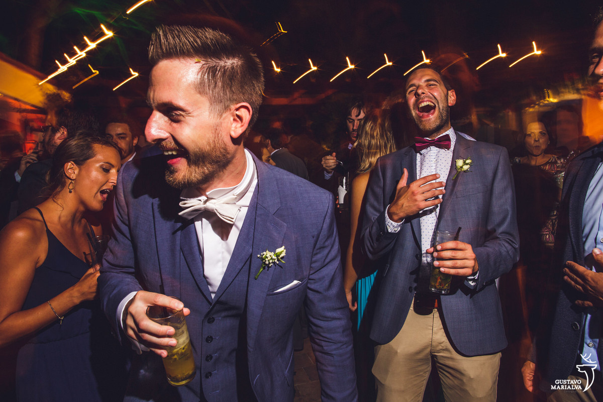 noivo dança sorrindo com o copo na mão enquanto amigo sorri ao fundo