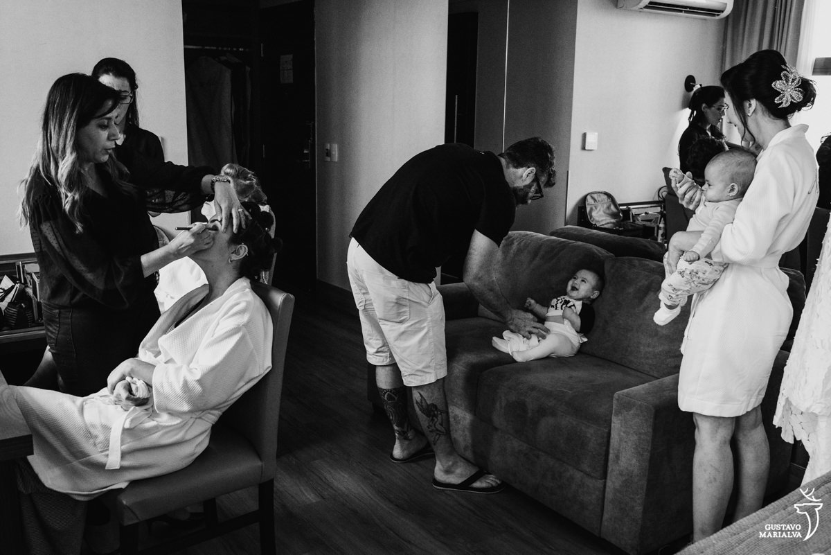 homem brinca com bebê no sofá enquanto mulher é maquiada durante o making of do casamento