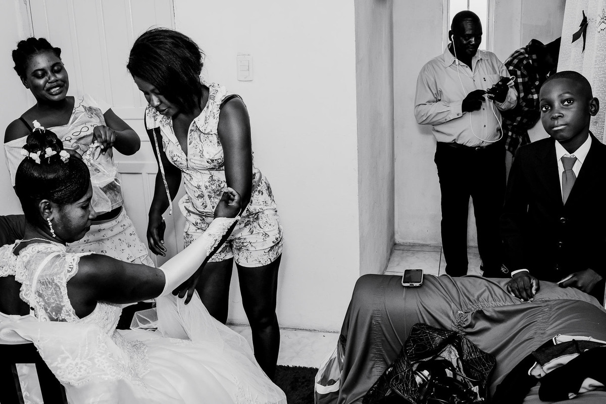 Na sala a noiva está se arrumando com a ajuda de duas amigas que estão lhe ajudando a fechar a luva. Perto do sofá uma criança haitiana encara a câmera, enquanto um adulto mexe no seu celular.