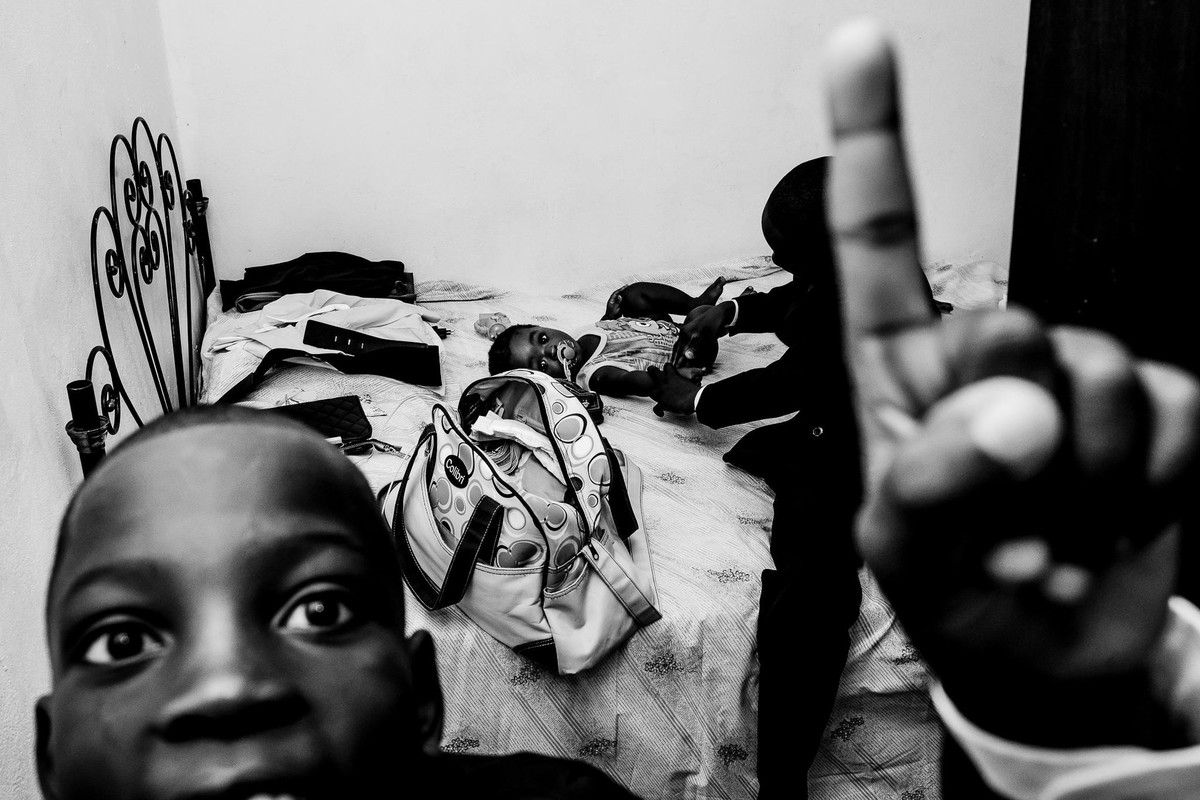Menino haitiano em primeiro plano levanta seu dedo olhando para a câmera enquanto atrás dele tem outro menino ajudando um bebê a se vestir sobre a cama