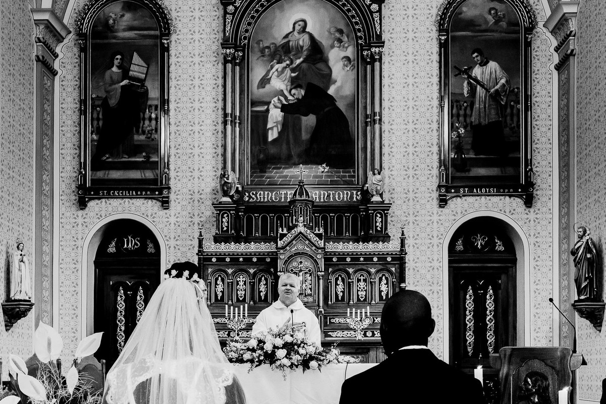 Os noivos estão lado a lado de costas para a câmera de frente para o altar da igreja matriz de Estrela decorada com painėis que compõem a cena em contraste com a pele branca do padre Azabido Ludwig
