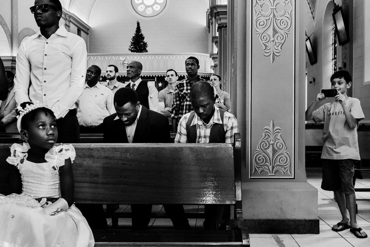 Enquanto o casamento haitiano está acontecendo é flagrada a presença de um menino branco registrando com seu celular numa das laterais da igreja enquanto aparecem demais haitianos sentados orando