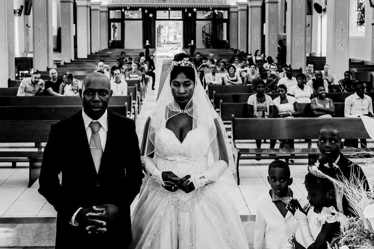 Os noivos haitianos estão em frente ao altar de pé cruzando as mãos em sinal de oração e ao seu lado estão os pajens e a daminha de honra e ao fundo os convidados sentados nos bancos da igreja