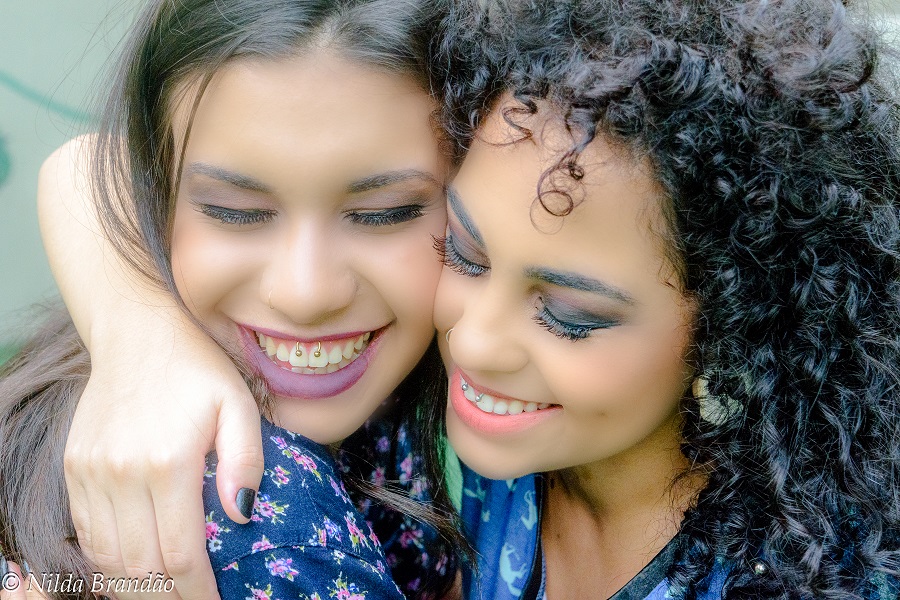 Amigas se abraçam, as duas usam piercing na gengiva