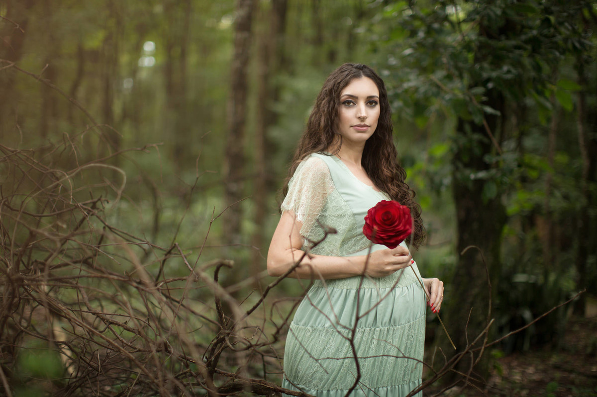 Grávida segurando flor vermelha na floresta verde. Fotografada pelo fotógrafo de gestante Rafael Ohana em Brasília-DF.