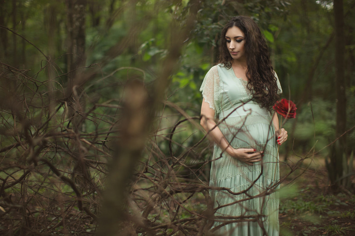 Grávida segurando flor vermelha na floresta verde. Fotografada pelo fotógrafo de gestante Rafael Ohana em Brasília-DF.