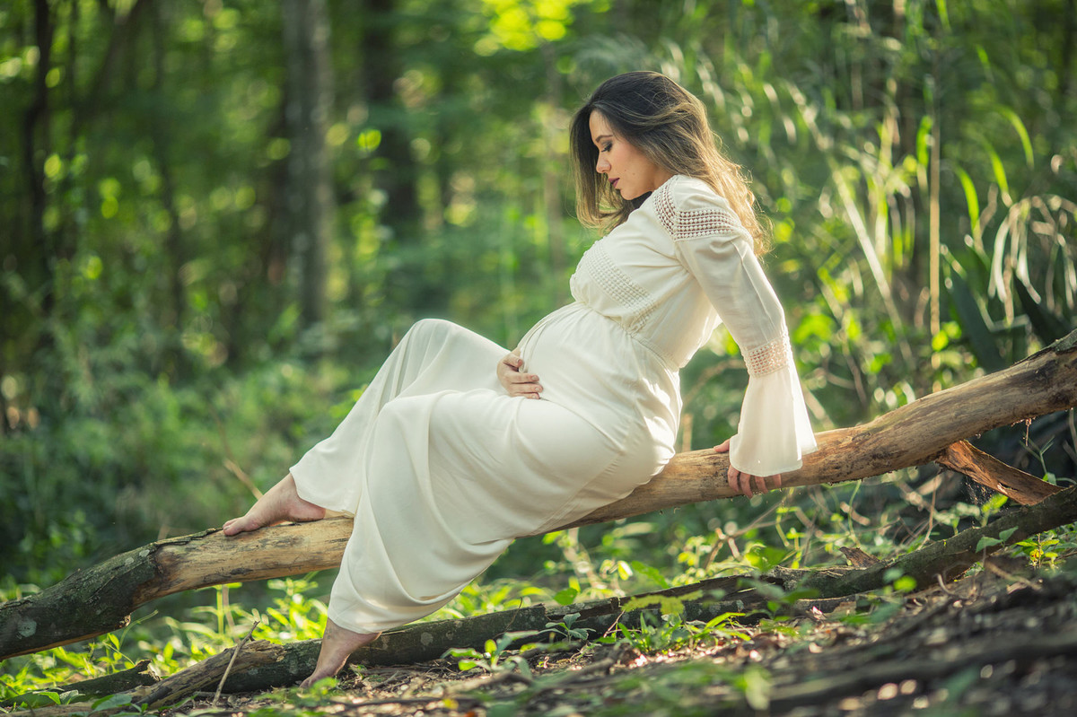 Gestante passando as mãos na barriga sentada no tronco de arvore. Foto feita pelo fotografo de gestante Rafael Ohana em Brasilia-DF