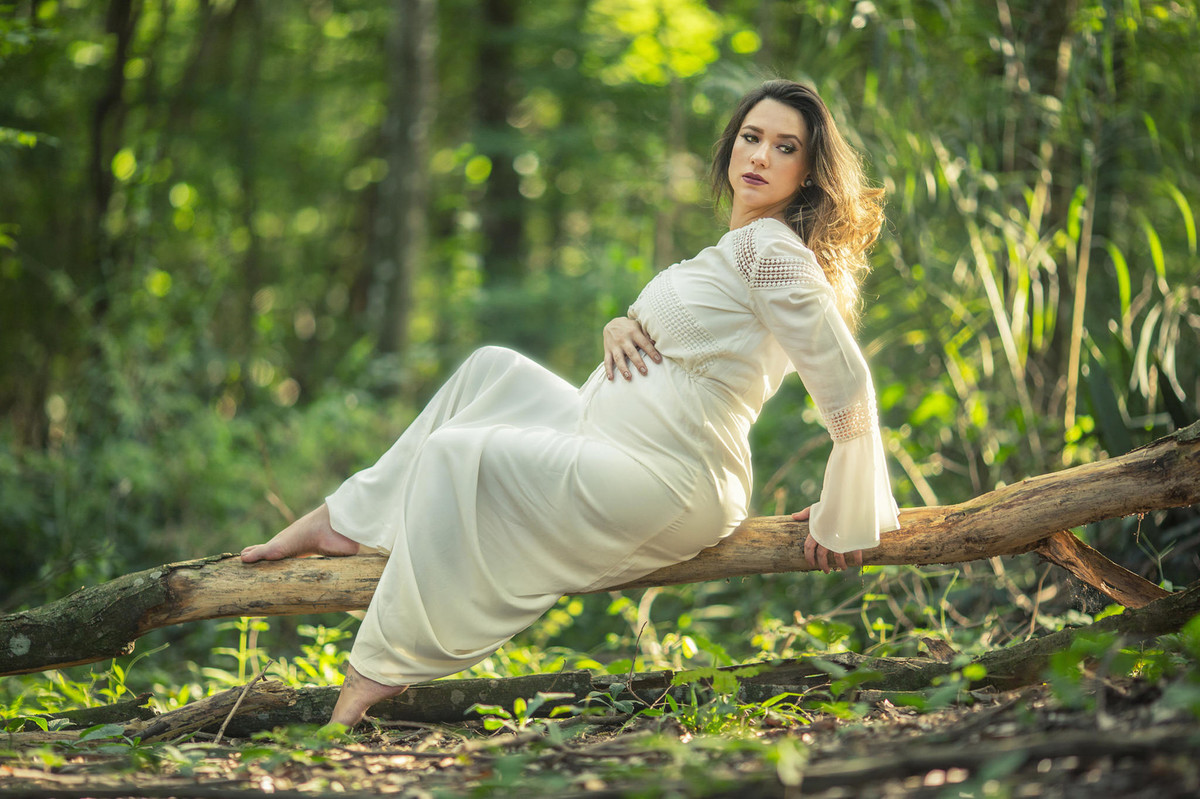 Gestante passeando com a mão na barriga sentada em arvore na floresta. Foto feita pelo fotografo de gestante Rafael Ohana em Brasilia-DF