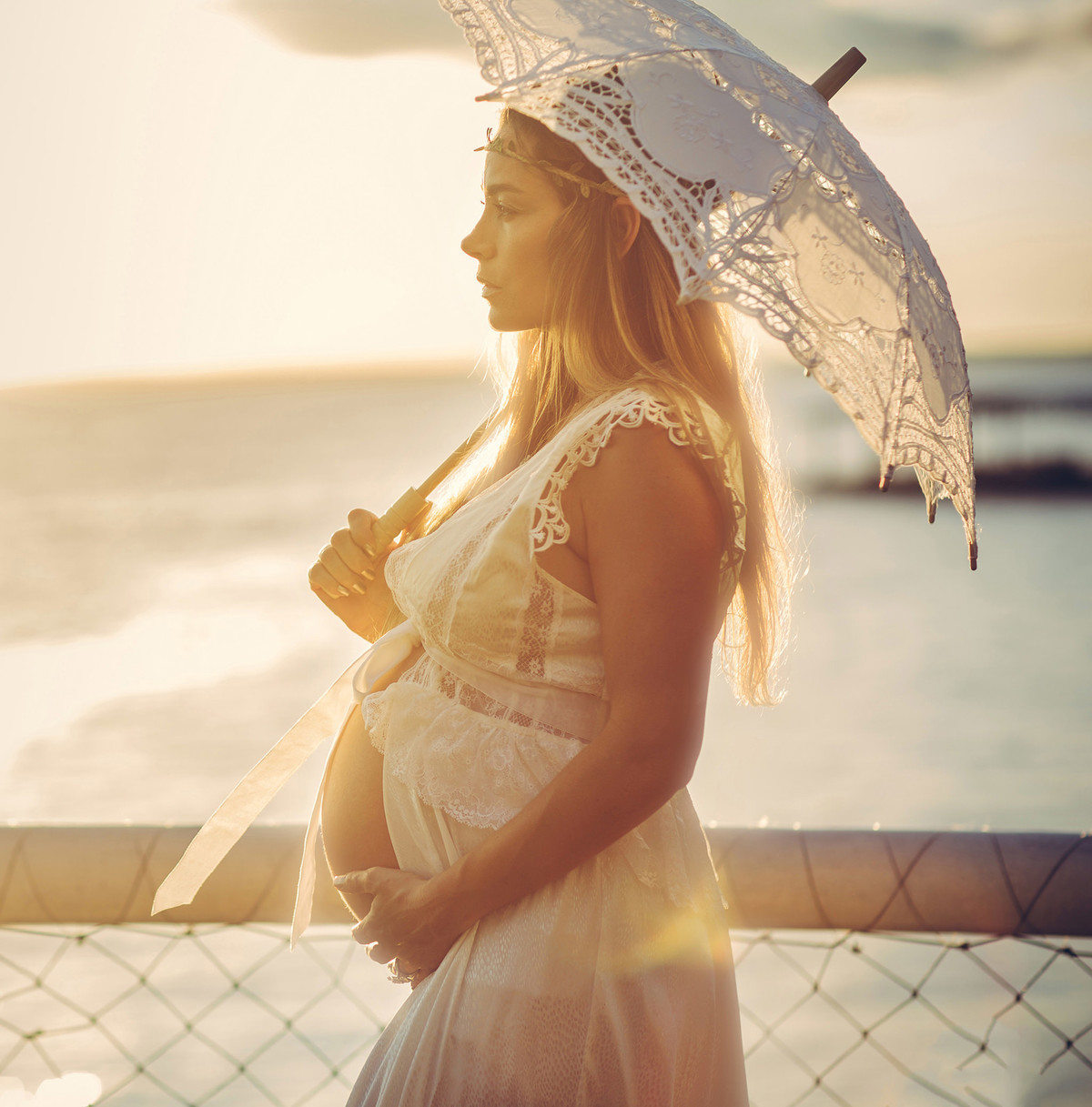 Grávida no por do sol usando vestido branco e utilizando sombrinha branca de renda. Fotografado pelo fotógrafo de gestantes Rafael Ohana em Manaus-AM