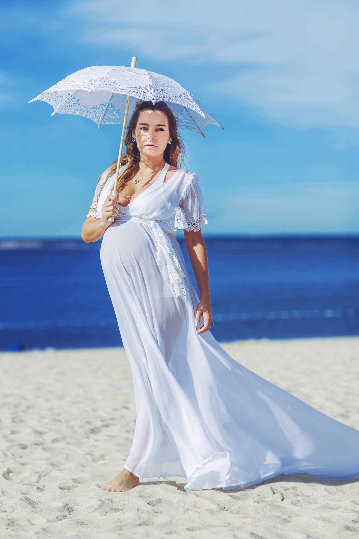 Grávida usando vestido branco e sombrinha branca na praia com areia branca. Fotografado pelo fotógrafo de gestantes Rafael Ohana em Manaus-AM
