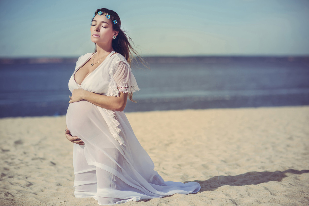 Grávida ajoelhada na areia segurando a barriga. Fotografado pelo fotógrafo de gestantes Rafael Ohana em Manaus-AM