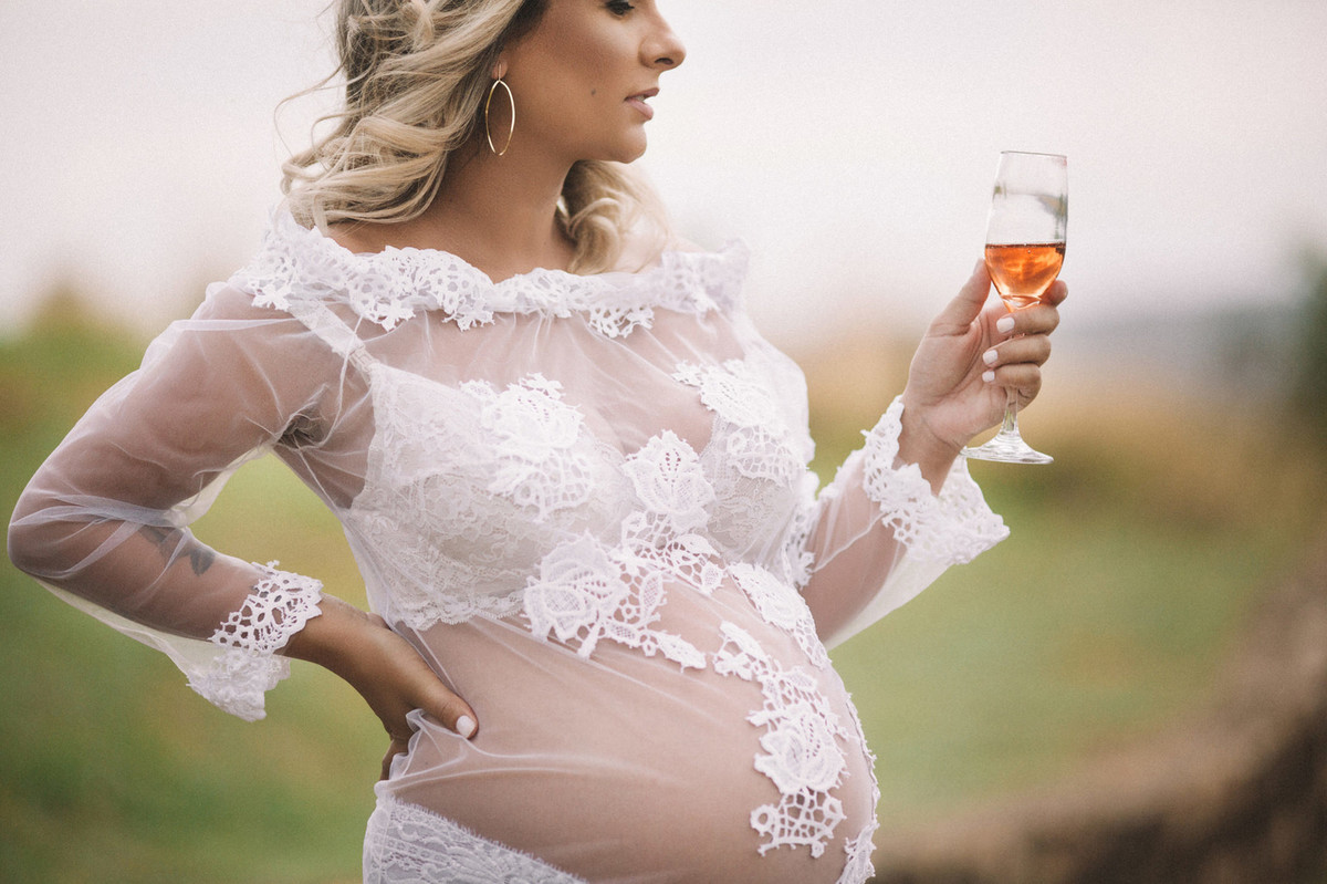 Gestante segurando uma tacinha de champagne usando vestido transparente. Clicado pelo fotógrafo de gestantes Rafael Ohana em Brasília-DF